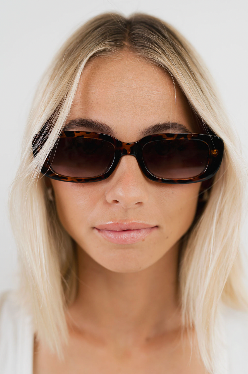 A close-up of a woman with blonde hair wearing oversized square tortoiseshell sunglasses with dark brown lenses, set against a plain white background.