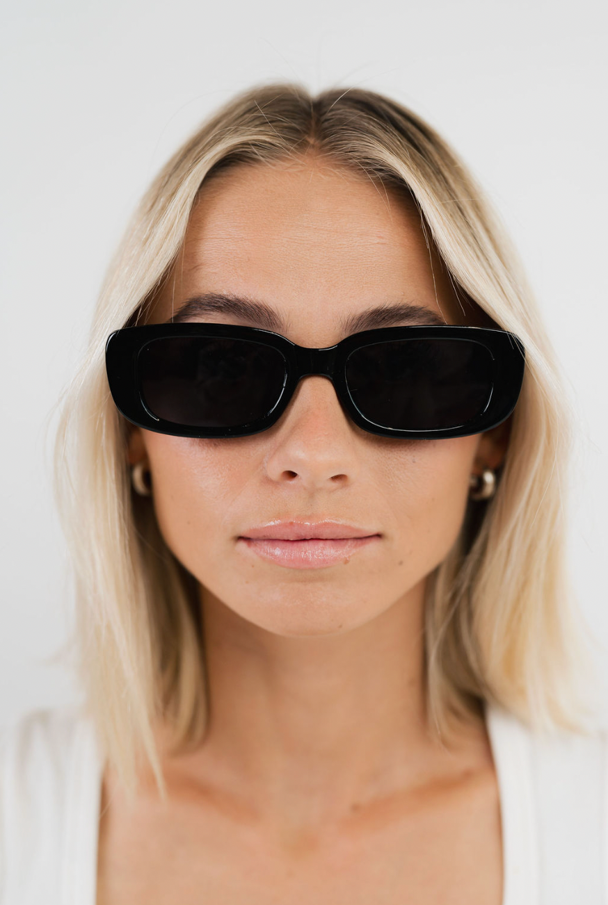 A close-up of a blonde woman wearing black rectangular sunglasses with dark lenses and a white top, set against a plain white background.