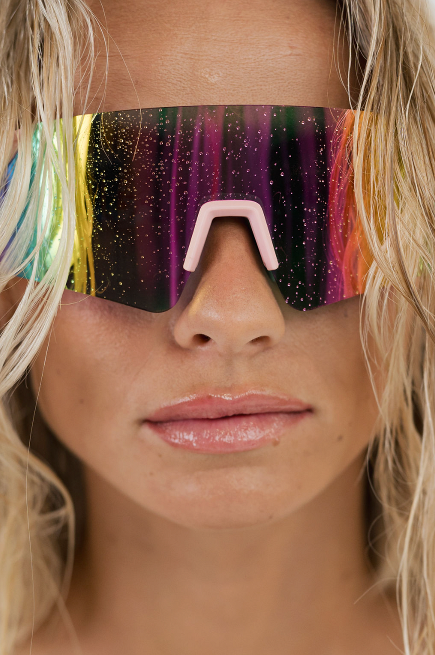 Close-up of a model wearing futuristic shield-style sunglasses with a colorful gradient reflective lens and a light pink nose bridge.