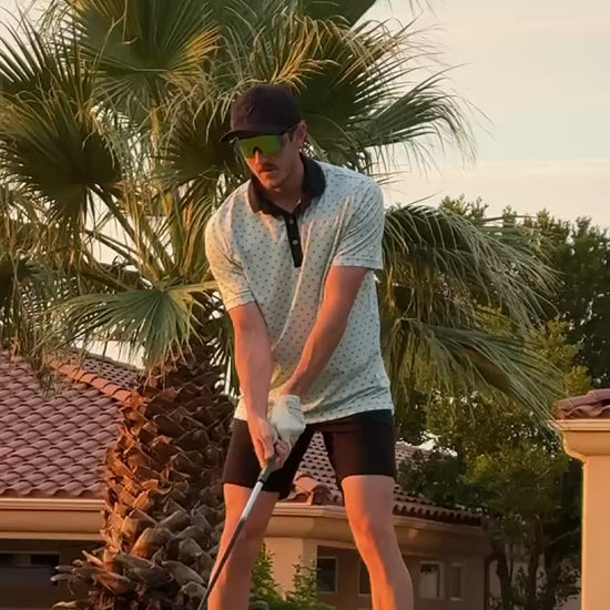 Golfer wearing shades of Rhae valocity sunglasses while hitting a golf ball on the grass with a palm tree in the background - Shades of Rhae