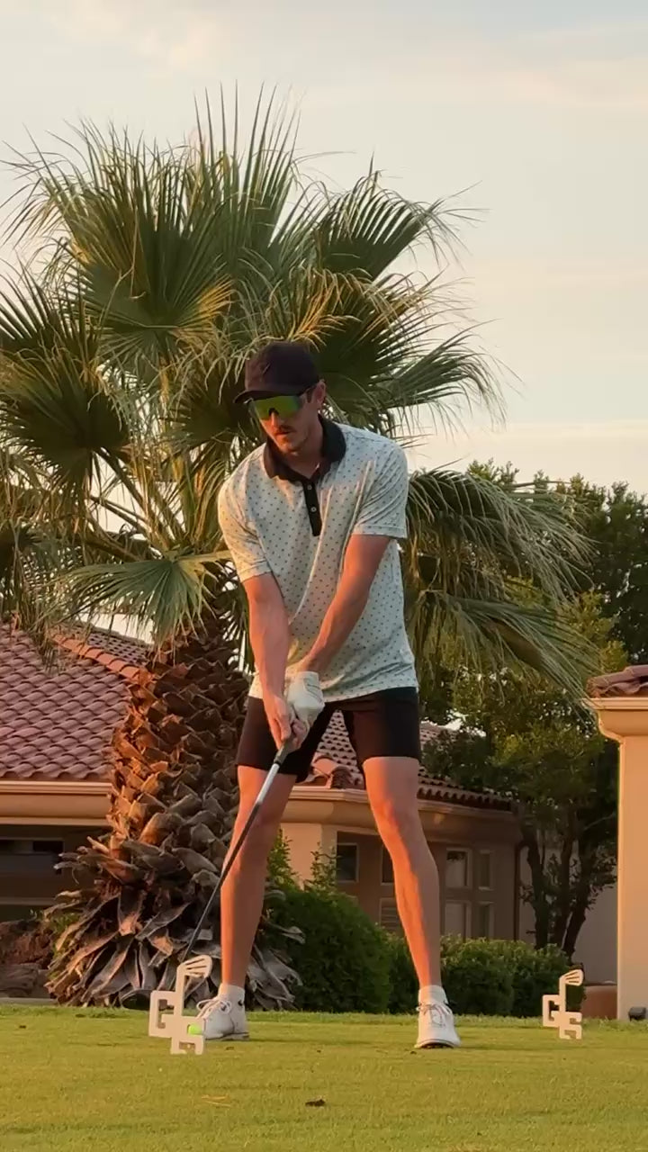 Golfer wearing shades of Rhae valocity sunglasses while hitting a golf ball on the grass with a palm tree in the background - Shades of Rhae